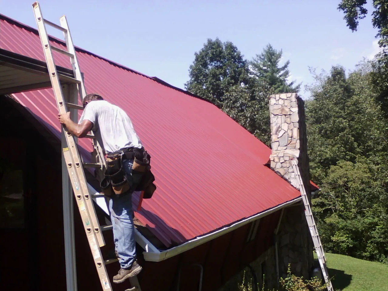 Roofer on a ladder beside a red metal roof and stone chimney — Soffit & Fascia Repair by Roofing Brothers in Garden Prairie